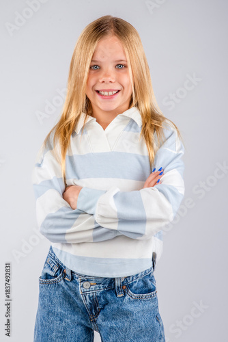 Cute smiling blond-haired girl standing with her arms crossed in a long striped polo shirt and blue jeans
