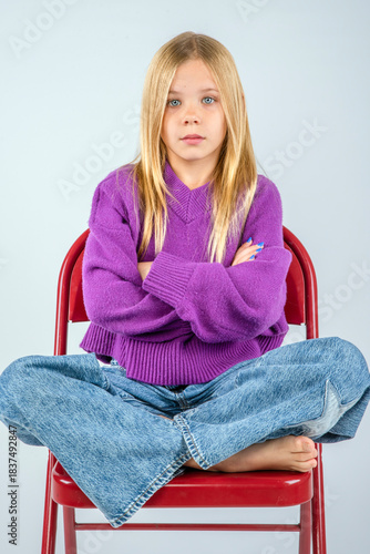 Young blond haired girl in a fuzzy purple sweater sitting with arms folded and legs crossed atop a red metal chair
