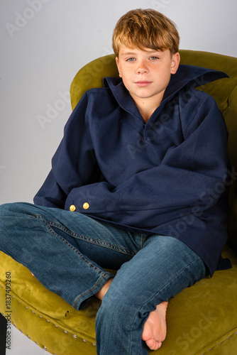 Handsome young barefoot boy in an oversized blue shirt, sitting casually in an upholstered chair, smiling and looking at the camera