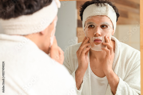 Quadro su tela Young man applying facial sheet mask near mirror in bathroom