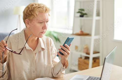 Focused woman with short blond hair sitting at desk with laptop, holding eyeglasses, looking at her smartphone. Frowning woman reading message, receiving unsatisfying news, having problems during work