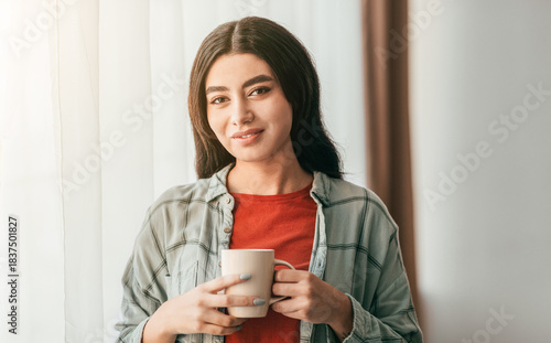 A young woman stands near a window, holding a cup. She has a warm smile and is clad in a cozy outfit. Sunlight filters softly through the curtains, illuminating her expression.