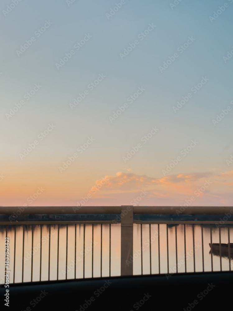 Obraz premium Sunset sky and calm river viewed through car window with bridge railing in foreground