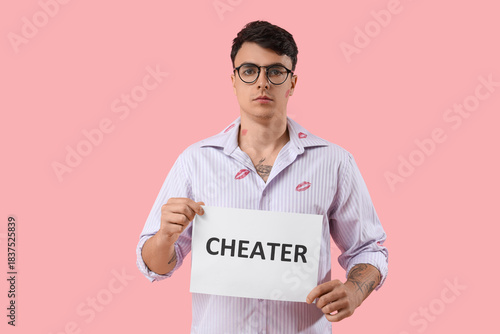 Фотография Young man with kiss marks on his shirt holding paper sheet with word CHEATER aga