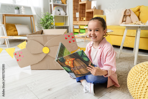 Cute little girl with cardboard plane reading adventure book at home