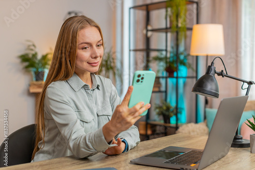 Happy mature middle-aged woman sitting at table desk using mobile phone smiling at home apartment. Girl freelancer texting share messages on smartphone social media applications online watching movie.