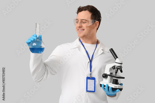Young male scientist with microscope and chemical flask on grey background