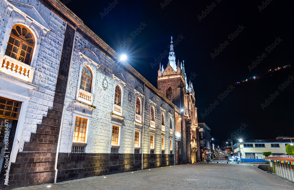 Obraz premium Illuminated Basilica of Our Lady of the Holy Water stands in Banos de Agua Santa, Ecuador. Semi-Gothic church features volcanic stone architecture and streetlights under night sky