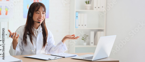 Female Asian doctor with headset and laptop giving online consultation in clinic
