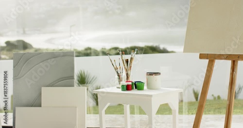 Camera is panning across stack canvases and easel with paint pots and brushes, outdoor view
