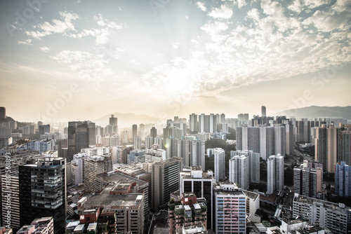 A complex urban skyline with varied skyscrapers under a partly cloudy sky, sun rays casting warm light over haze-covered mountains.
