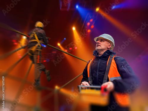 Stage construction crew installs high-level rigging under concert lighting, ensuring safe setup of event structures and coordinating technical assembly tasks.