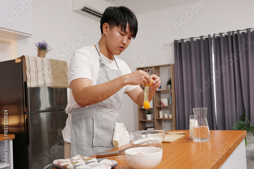 Asian young man cracking egg into bowl while cooking in modern kitchen, Asian male, apron, countertop, whisk, mixing bowl, milk jug