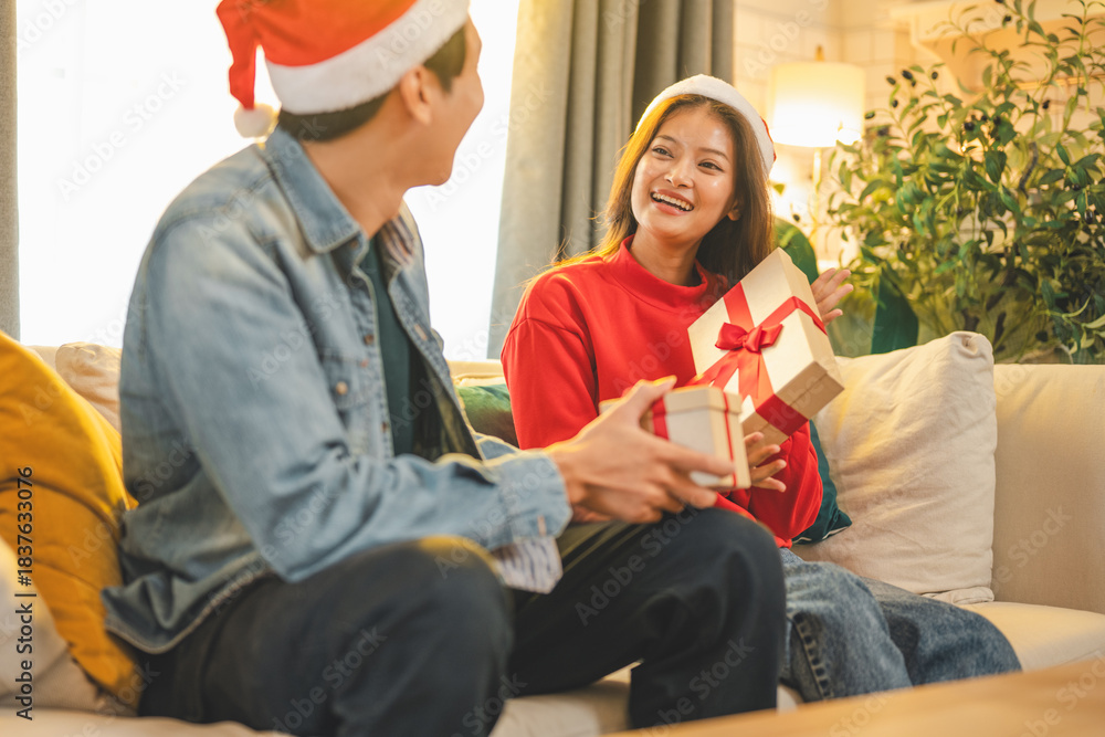 Naklejka premium Two young people enjoying a cheerful Christmas moment together at home, exchanging gifts with smiles. The warm festive scene highlights joy, celebration, and the cozy spirit of the holiday season.