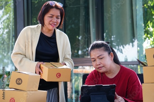 Two women are working outdoors, with one standing and holding a package box while the other sits looking at a tablet.