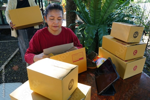 A woman is packing an item into a package box surrounded by a tablet and other boxes, with another woman holding a box behind her.