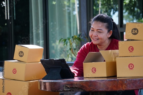 The woman is smiling while looking at a tablet and working with package boxes stacked on a table.