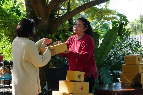 Two women stand and hand a package box to each other, with other boxes stacked around them.