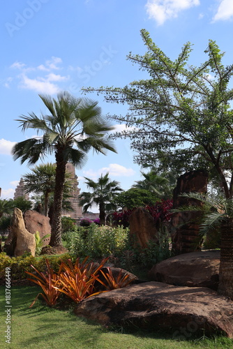 An outdoor setting with a small, well maintained garden in the foreground. A variety of tropical plants can be seen, with one particularly large palm tree standing prominently