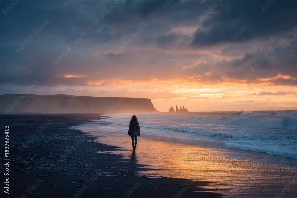 Fototapeta premium Woman strolls along a black sand beach in Iceland during a vibrant sunset with waves gently crashing in the background