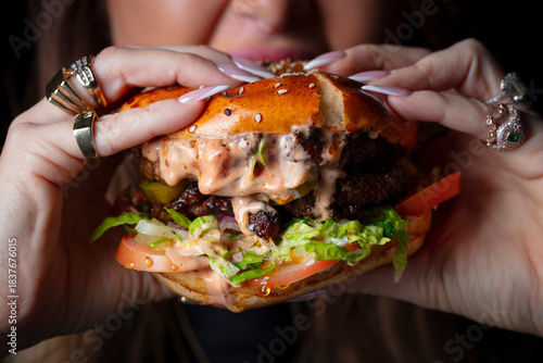 Fotografie A woman biting into a thick hamburger with Thousand Island dressing, lettuce, and tomatoes
