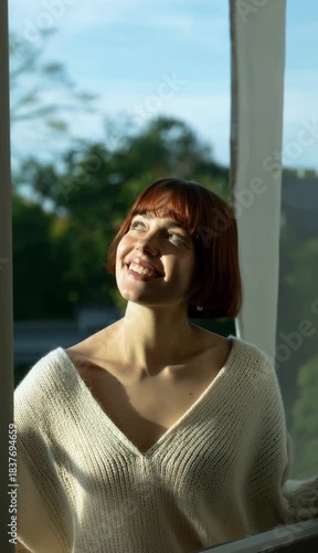 Young Woman Smiling Brightly Near Window in Natural Light with Short Red Hair and Casual Sweater