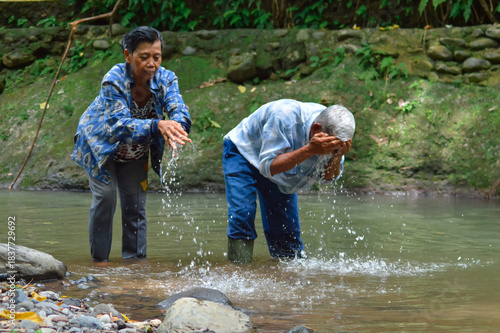 An elderly couple stands in a shallow stream, the man in a plaid shirt splashes water on his face, while his wife in a blue jacket washes her hands nearby in the lush green environment.