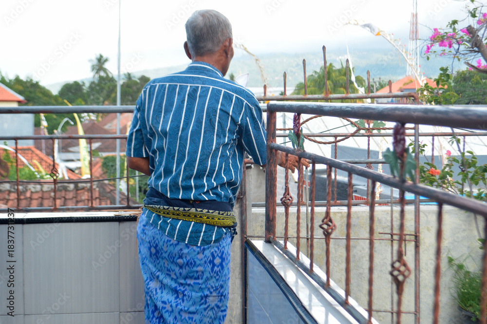 Obraz premium A back view of an elderly man in a blue sarong and yellow sash standing on a balcony, gazing over a village landscape with hills and clear sky in the background.