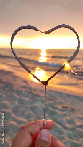 Burning sparkler in shape of heart against backdrop of sea at sunrise. Heart shaped sparkler burning on sea during sunrise sunset. Silhouette of heart in woman hand on seashore. Sparks fire. Vertical