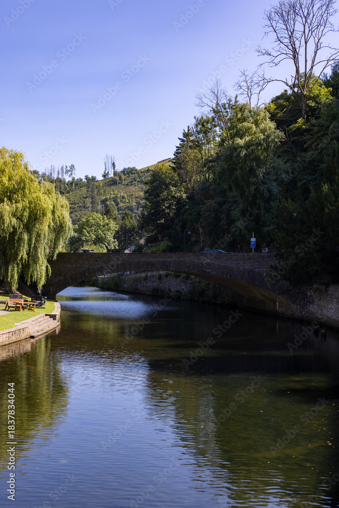 Fototapeta premium Esch-sur-Sure, Wiltz, Grand-Duche de Luxembourg, September 07, 2025, Gentle footpath alongside calm flowing stream, Peaceful pathway beside gentle river bend with trees and arch reflection