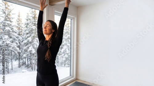 Calm woman in black sportswear practicing yoga with arms raised, stretching at home by a window with a snowy winter forest view.