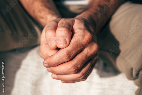 Closeup of clasped hands of a mature man sitting indoors, showing tension, stress, and emotional struggle.