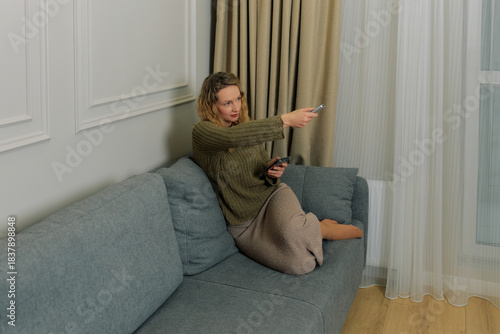 Woman watching TV on a couch and pointing remote control at screen while relaxing at home in the evening