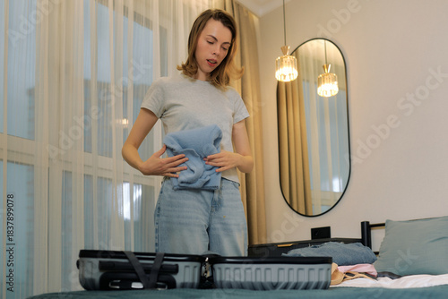 Woman packs clothes into suitcase in modern bedroom with mirror and light fixtures
