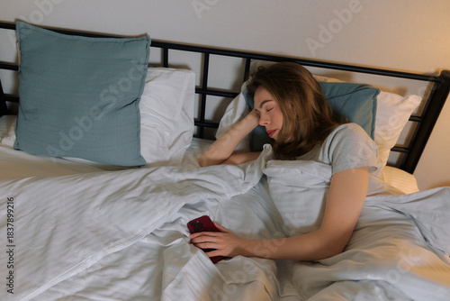 Woman sleeps in bed holding smartphone with soft lighting in a cozy bedroom at night