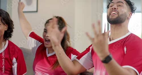 Diverse friends wearing red-white jerseys on couch watching goal, pumping fists and discussing play
