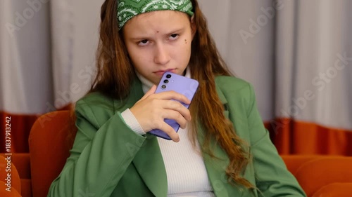 Teenage girl with green bandana and long hair looks at her phone with a tense face, reading upsetting chat messages and thinking about how to respond, concept social media drama and online bullying.