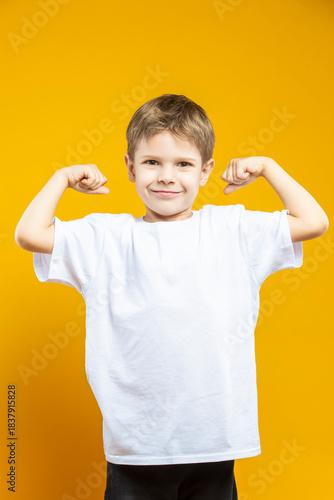 Smiling Male Kid Boy Of 5-6 Years Old Wearing White t-shirt Polo With Lifted Hands As Power Gesture Isolated on Yellow Background As Children Indoor Portrait To Convey Childhood Lifestyle Concept