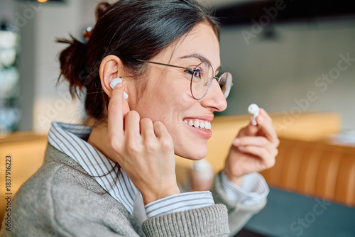 Young woman smiling, wearing glasses, inserting wireless earbuds into her ear, enjoying audio content. Modern technology