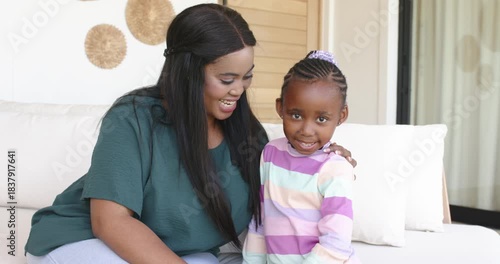 African American mother and daughter smiling at camera sharing gentle touches and giggling on sofa