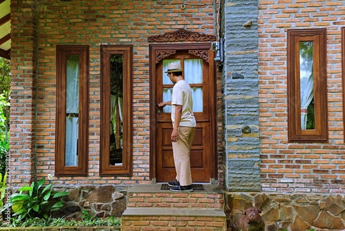 A man facing a wooden door on a rustic brick house exterior in daylight.