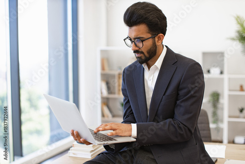 An Indian businessman wearing glasses works on his laptop in a modern office setting