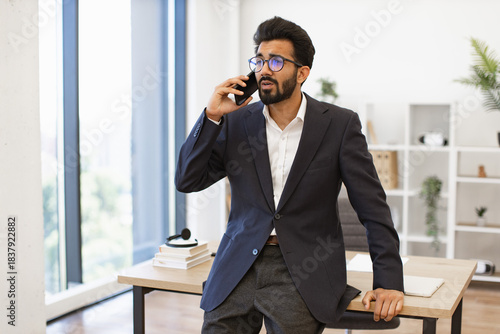 An Indian businessman wearing glasses is talking on the phone in his office, looking concerned