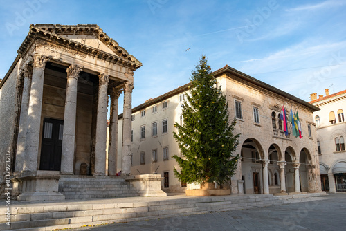 Wide view of Pula’s Forum square featuring the Temple of Augustus and a tall decorated Christmas tree under clear blue sky. 