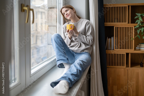Photos Happy dreamy woman enjoys quiet time pause holding coffee mug relaxing sits on windowsill