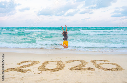 New Year 2026 written on sand, happy woman with hands up jumping on the beach