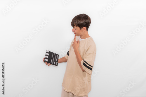 young casual dressed Generation Z influencer white male model teenage boy considering how to create and direct social media film reel with blank clapperboard slate. plain background studio setting