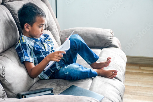 Side View Of Focused Young Asian Boy Studying And Reading Notebook On Sofa