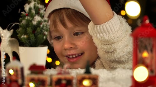 Close up of little girl in santa hat sprinkles snow on toy fir on background of Christmas tree. Happy child plays with New Years gift