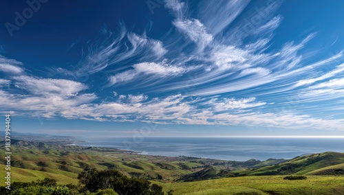Verdant hills roll toward the vast ocean under a bright, blue sky filled with streaky clouds